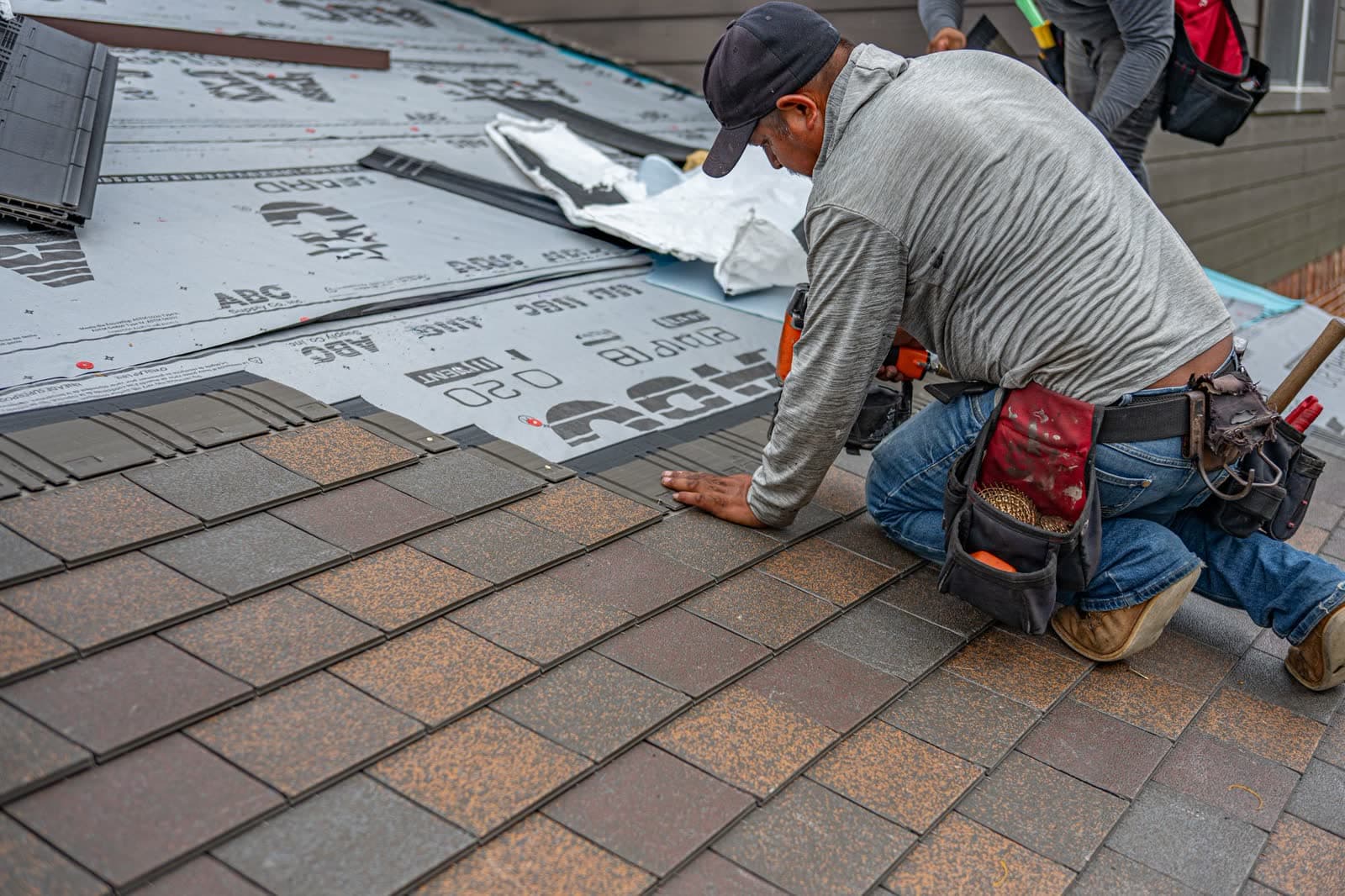 A roofer in a gray shirt and cap kneels on asphalt shingles and fastens them in daylight.