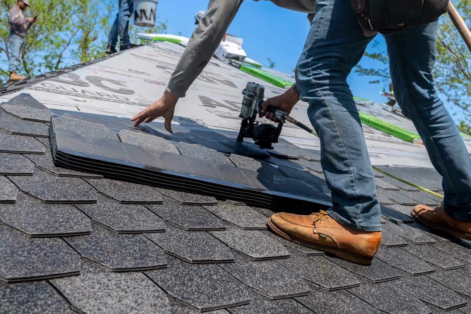 A roofer drives a nail gun into asphalt shingles on a sloped roof.