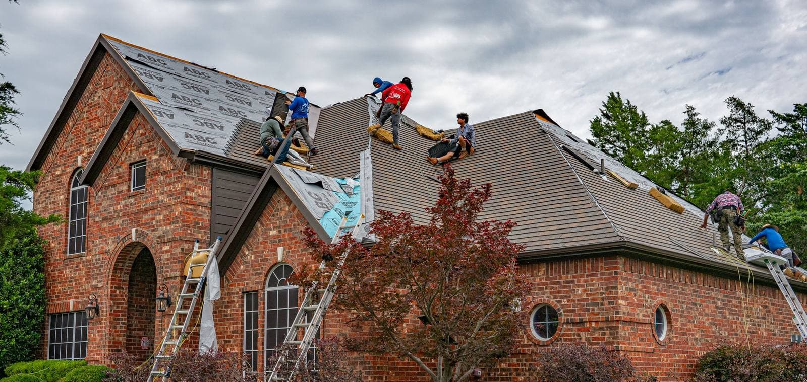 A roofing crew tears off and re-shingles the roof of a brick house, with ladders against the walls on an overcast day.
