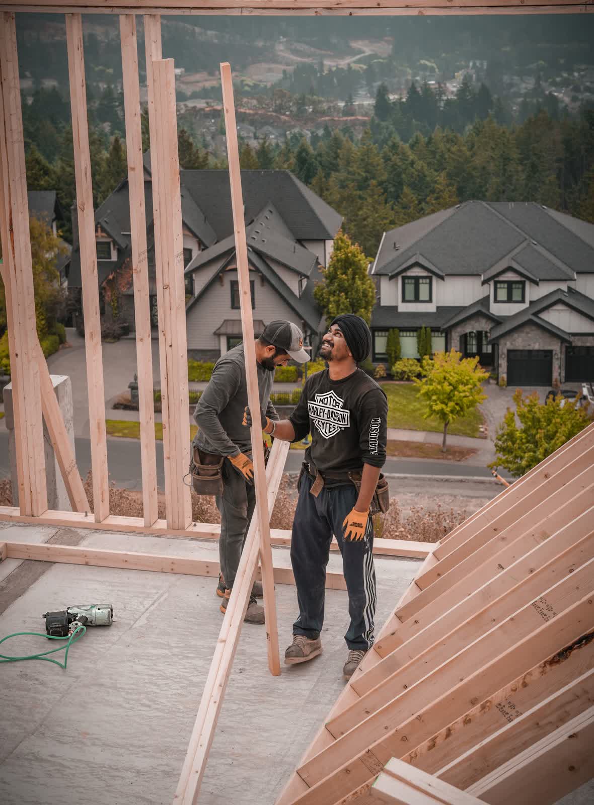 Two workers handle lumber while assembling a house frame on a construction site.